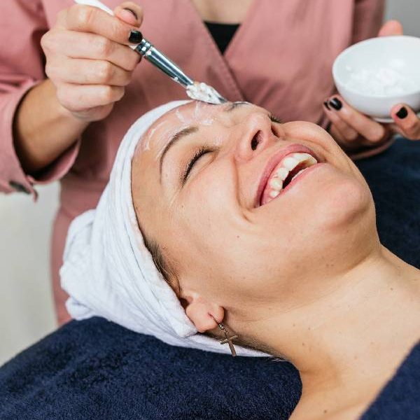 Mujer sonriente recibiendo un tratamiento facial en un spa, mientras otra persona aplica una mascarilla con brocha. La mujer está relajada, con una toalla en la cabeza y rodeada de toallas azules.