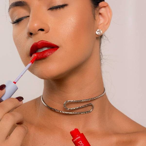 Mujer aplicando labial rojo con un aplicador, luciendo un collar plateado y un maquillaje radiante.