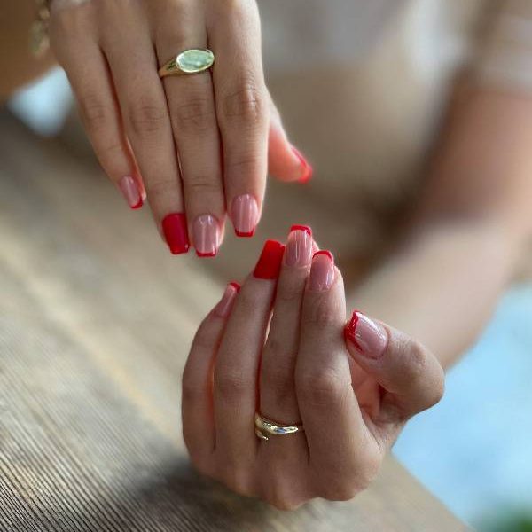 Manos con uñas manicuras en tonos rojos y rosas, destacando un diseño moderno. Se observan anillos dorados en los dedos, sobre una superficie de madera.
